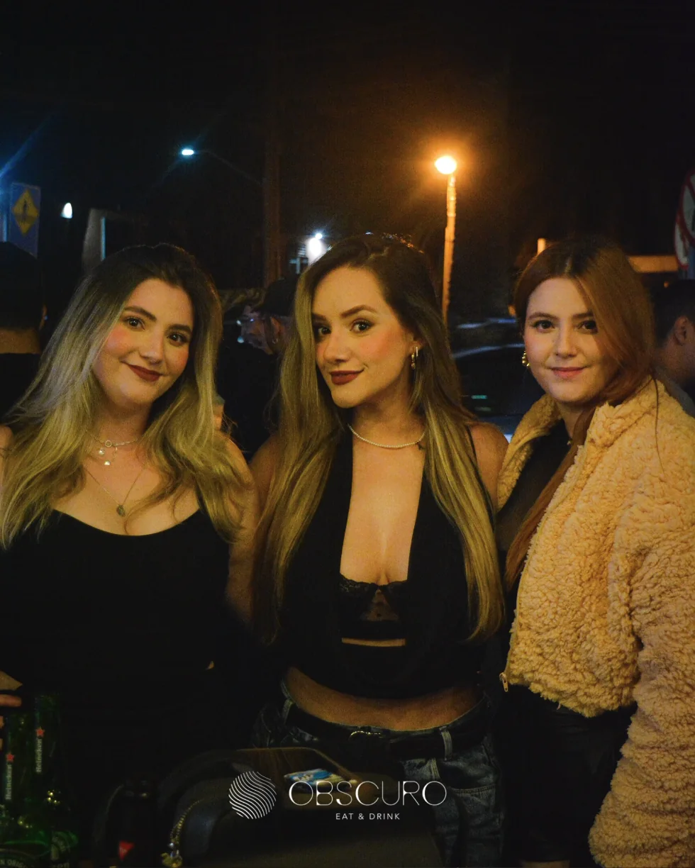 Três amigas posando à noite na frente do Obscuro, iluminadas pela rua