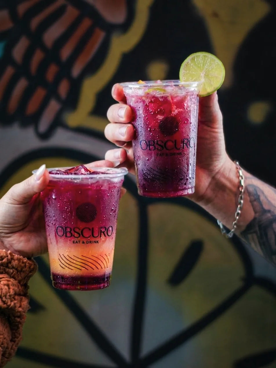 Duas mãos brindando com drinks roxos em copos do Obscuro, em frente a um mural grafitado